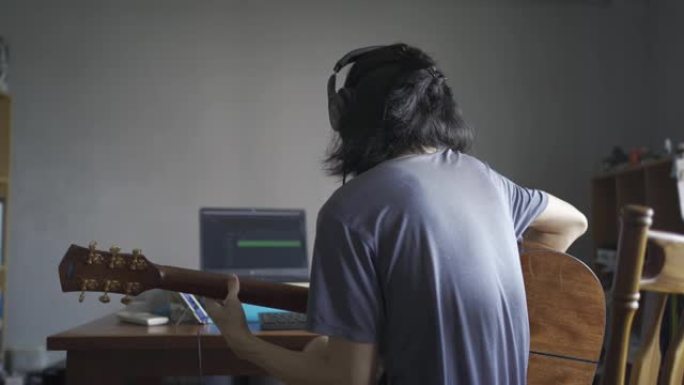 一个留着长发的男人戴着耳机，弹木吉他，以便在家庭房间里录音到计算机中