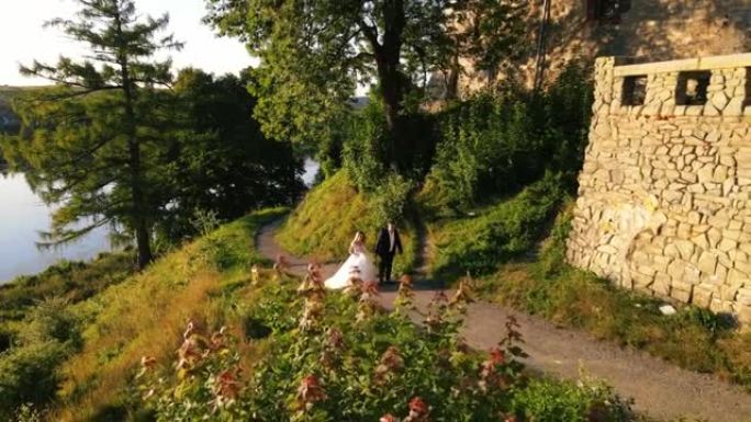 新婚夫妇手牵手走在古老城堡和湖泊附近的绿色公园里。一对年轻夫妇在日落时走在公园的小径上。新娘拿着礼服