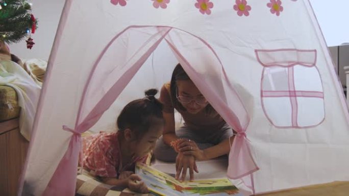 亚洲妈妈正在给她可爱的小女孩讲一本书作为睡前故事，她躺在家里可爱的帐篷里的地板上，享受着快乐和快乐，