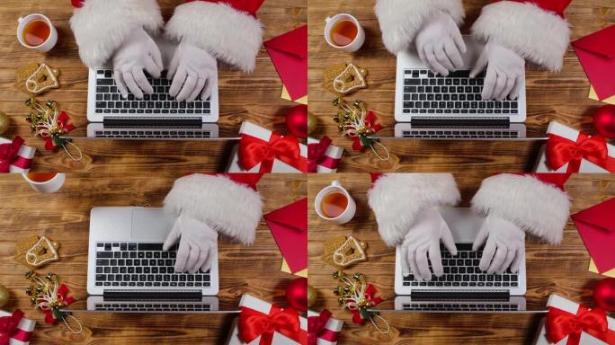 顶视图圣诞老人手在键盘上打字，并在木制装饰的桌子上喝茶。圣诞老人与笔记本电脑一起工作，浏览孩子们的来