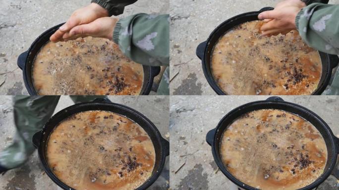 一名男子厨师用手掌摩擦孜然，然后将调味料倒入一个大锅中，并加热抓饭。露天烹制乌兹别克国菜。慢动作，特
