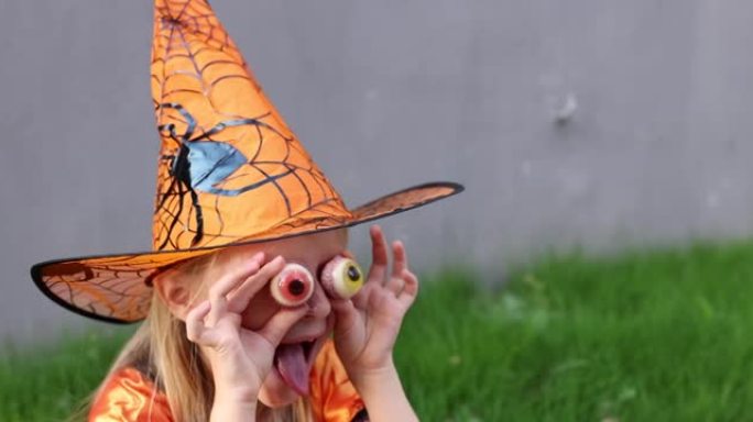 可爱的金发小白人女孩，七岁，穿着帽子和黑色橙色连衣裙的女巫服装，在街上户外庆祝万圣节。假日概念。慢动