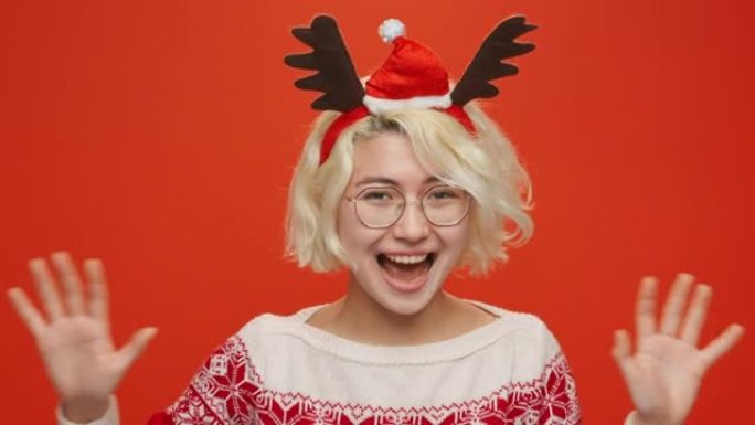 圣诞贺卡有趣的年轻亚洲女子出人意料地出现在圣诞服装的框架中，鹿角微笑着看着红色背景上的相机。节日气氛