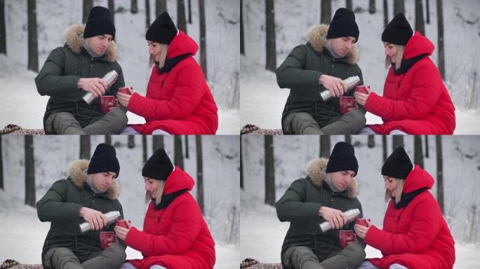 恋爱中的年轻夫妇坐在冬季森林里从热水瓶里倒热茶。