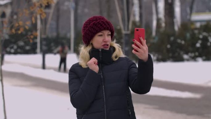 一个冬天白雪皑皑的公园里的女人拿着智能手机，通过视频通话。女孩向对话者打招呼，显示拇指向上的手势，发