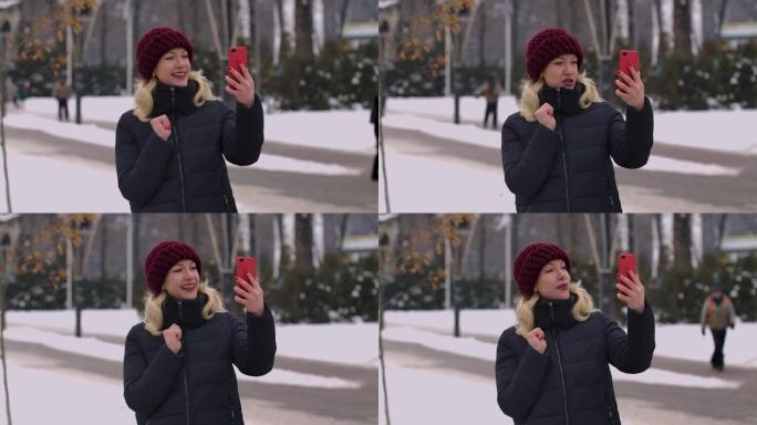 一个冬天白雪皑皑的公园里的女人拿着智能手机，通过视频通话。女孩向对话者打招呼，显示拇指向上的手势，发