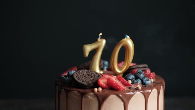 在黑色背景的巧克力生日蛋糕上吹蜡烛，搭配70号金色蜡烛。70周年生日蛋糕。慢动作