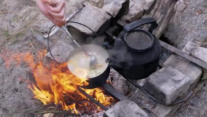 男性在户外树林里的篝火上的平底锅里准备食物。缩放。特写