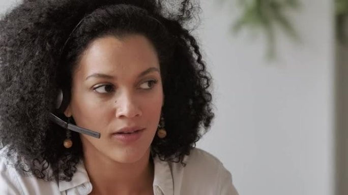 使用耳机在视频电话会议上交谈的年轻非洲裔美国妇女的肖像。穿着白色上衣卷发的黑发女人坐在家庭办公室的桌
