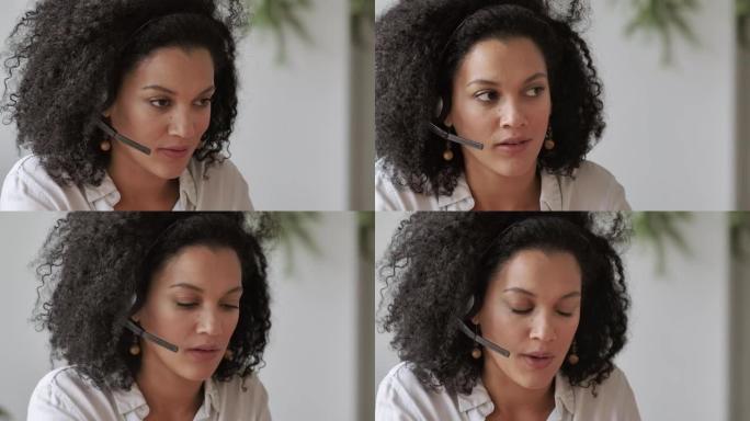 使用耳机在视频电话会议上交谈的年轻非洲裔美国妇女的肖像。穿着白色上衣卷发的黑发女人坐在家庭办公室的桌