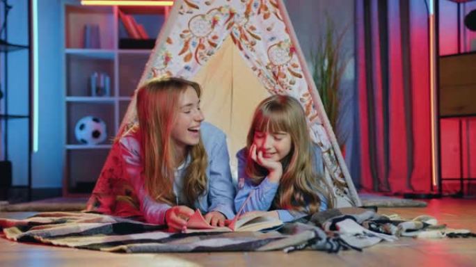 可爱的微笑快乐友好的两个不同年龄的姐妹在装饰帐篷的床罩上放松，谈论他们晚上读的书