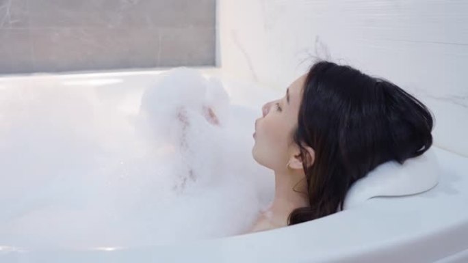 特写开朗的亚洲女人在浴缸里吹泡沫