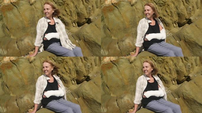 资深微笑的老年妇女在大风天坐在岩石海岸线上放松，患有关节疼痛。手臂受伤。老人手腕在石膏吊带绷带的感觉