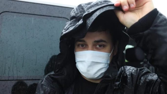 一个穿着黑色外套的男人在大雨中，脸上戴着医用口罩。他淋湿了，环顾四周。
