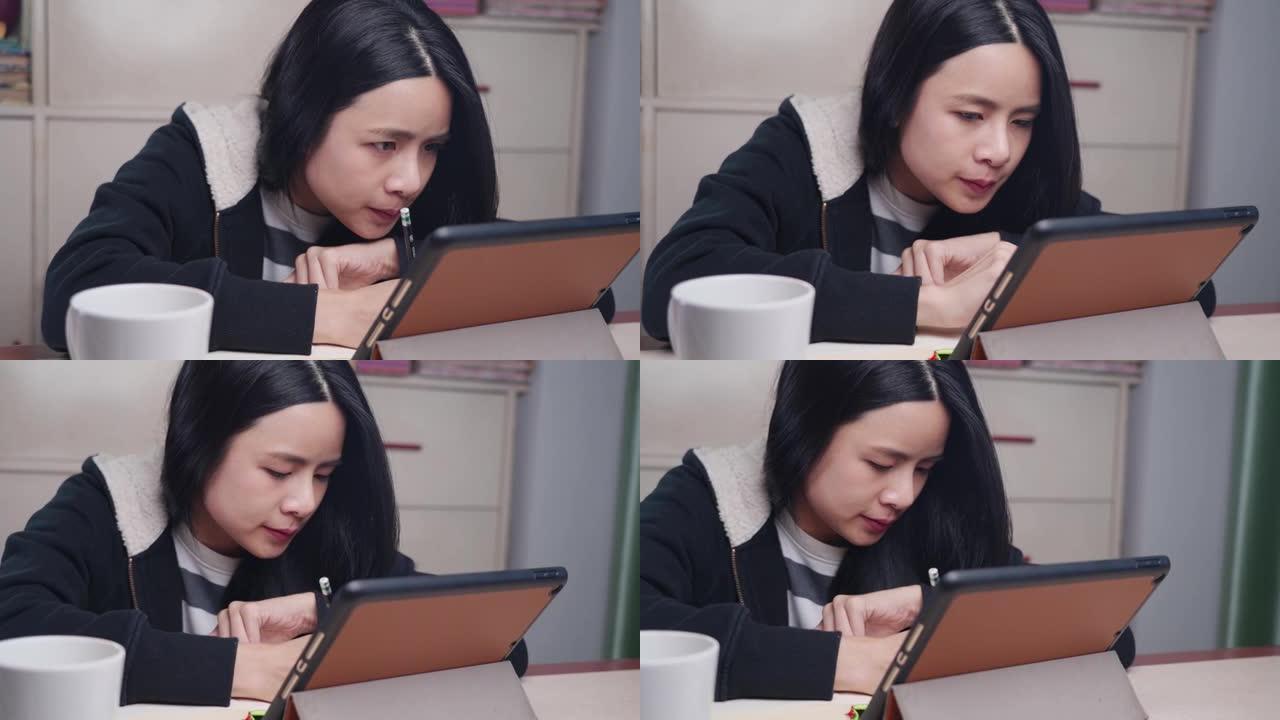 年轻的亚洲女孩看平板电脑在线学习课，用铅笔在笔记本上写下，专注于家庭作业，疏远学费，家庭学校，在家工