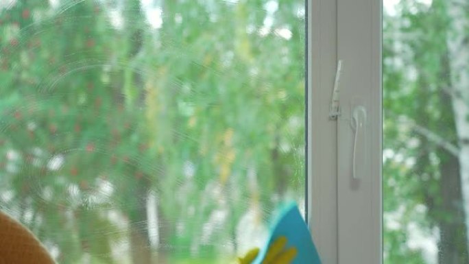 夏天洗塑料窗。一名妇女的手戴着黄色橡胶手套，用清洁剂，蓝色抹布和刮刀清洗窗户