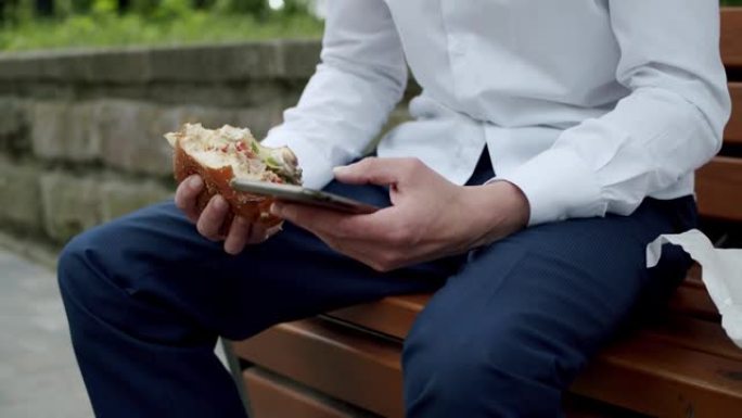 坐在街头长凳上的男子在智能手机上打字并拿着汉堡