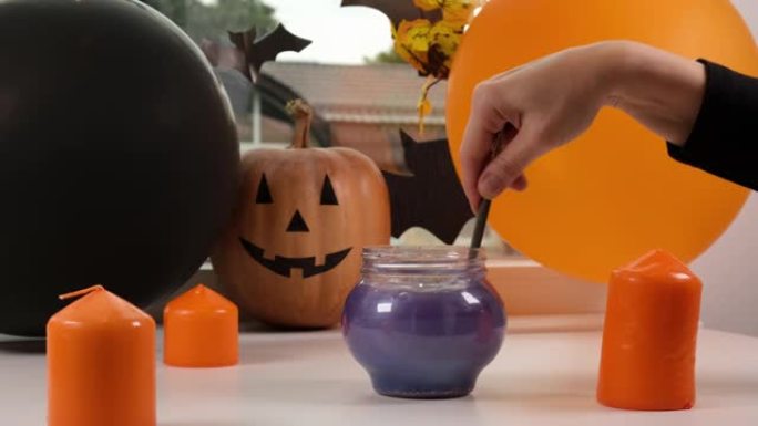 万圣节南瓜、蜡烛和气球。女巫正在制造魔法药水。女人的手在杯子里搅拌柔软的紫色物质