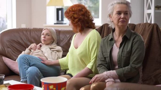 红发成年女性的肖像与坐在客厅触摸肩膀的朋友争论，看着相机摇头。在室内与得罪的朋友在一起使白人女士心烦