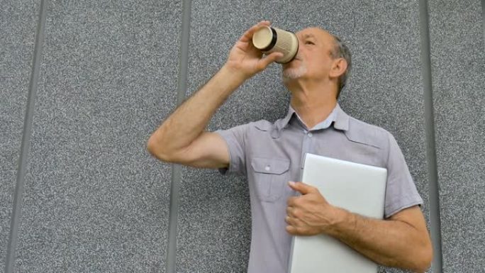 年长同事的男性肖像，带着银色的笔记本电脑和一杯咖啡，早上在深灰色的墙壁附近等待会议，远程工作，喝咖啡