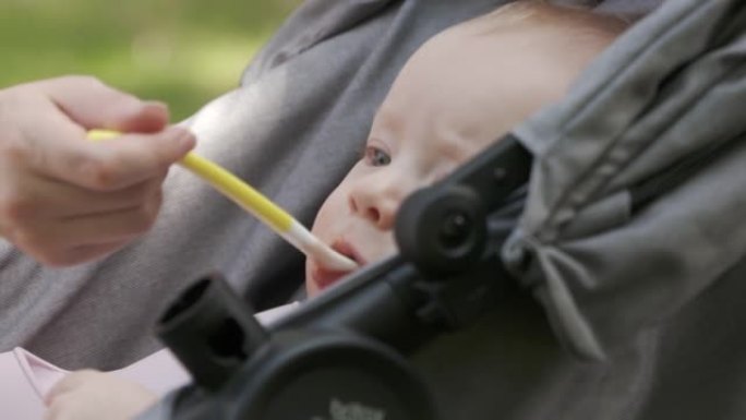 可爱的小男孩坐在婴儿车里，和妈妈一起在户外散步，吃勺子里的第一份食物，妈妈给儿子喂泥食物。