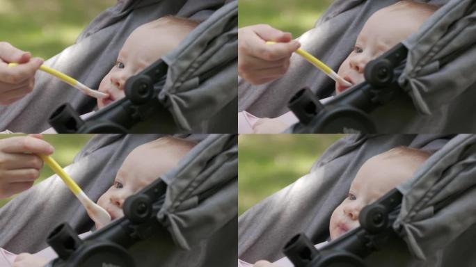 可爱的小男孩坐在婴儿车里，和妈妈一起在户外散步，吃勺子里的第一份食物，妈妈给儿子喂泥食物。