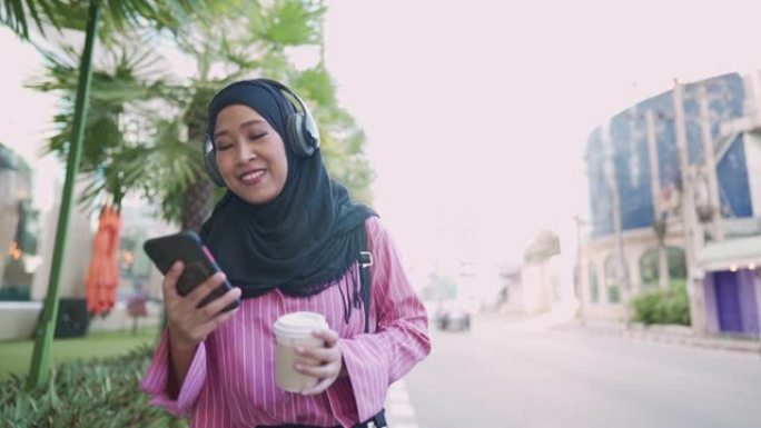 年轻的亚洲穆斯林女性戴着耳机听播客，用智能手机拿着咖啡杯走在街边的下午咖啡，享受音乐和户外，办公室工