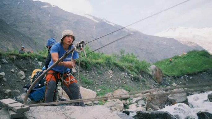 一名男子拉着一根绳子，把背包系在卡宾枪上，越过一条山间的河流。