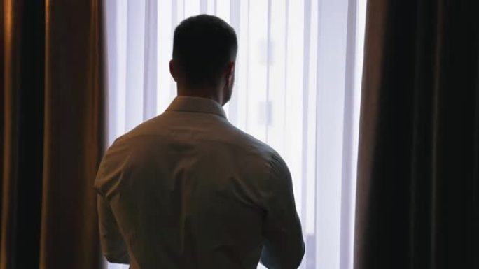 酒店里窗户附近的人。穿着白衬衫的年轻商人站在窗户附近，在酒店房间里透过窗户观看。后视图。