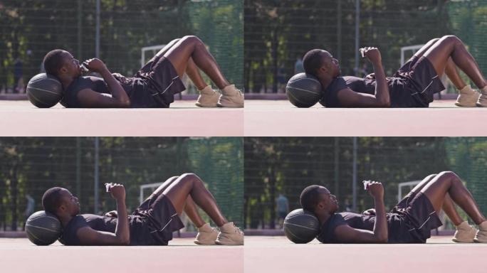 年轻疲惫的非洲裔美国人在街球游乐场的地板上休息，躺在篮球上，吃蛋白质棒