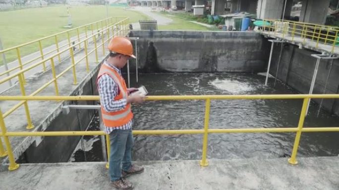穿着反光背心和头盔的亚洲男性工程师在工厂检查废水处理池。使用数字平板电脑的人