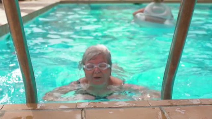 灰白头发的老年妇女在酒店游泳池游泳，眼睛上有谷歌，靠近时微笑着满足