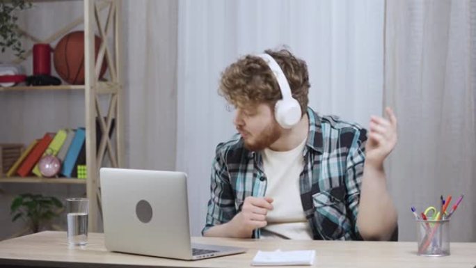 穿着方格衬衫的年轻人喜欢戴着白色大耳机的音乐。红发男性，留着胡须，坐在家庭办公室靠近笔记本电脑的桌子