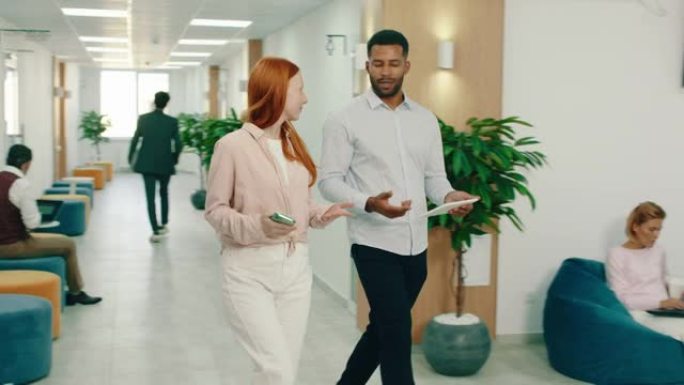 一个黑人正在办公室里和一个漂亮的姜黄色女人一起散步，他们坐在办公室的休息区，身后有三个人在咖啡桌旁工