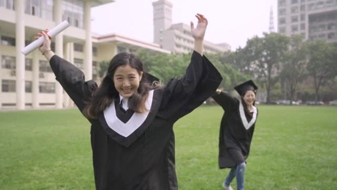 两个兴奋的快乐的亚洲女学生站在地幔的大学礼堂里，手里拿着文凭微笑着，脸看着相机。自信的应届毕业生双臂