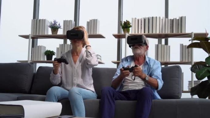 老年成熟夫妇玩虚拟现实眼镜玩得开心，老年夫妇坐在沙发上拿着摇杆边笑边玩电子游戏，快乐老年科技概念