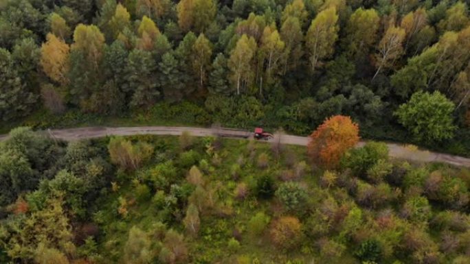 鸟瞰式农用拖拉机沿着乡村道路驶过秋天的森林。农用拖拉机、农用机械农业自然季节性工程。空中无人机视图秋