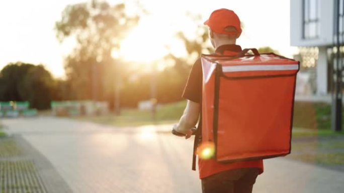 年轻的快递员骑着他的电动滑板车。交付男性包裹递送服务人员骑着电动滑板车在日落时带着红色背包在街上的空