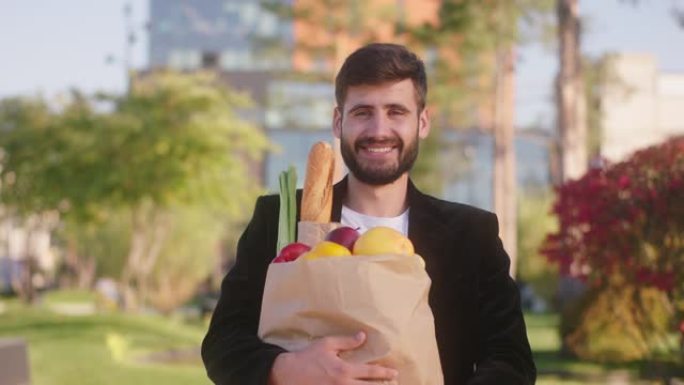在一个阳光明媚的日子里，笑容灿烂的帅哥在现代街道中间的镜头前摆着一个装有食物的袋子摆姿势