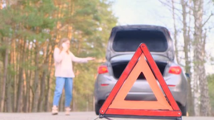 汽车故障。一名年轻女子沿着树林的道路走，旁边是一辆破汽车在打电话。紧急三角特写，一个离焦的女人。汽车