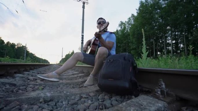 戴着墨镜和帽子的男人坐在铁路铁轨上弹吉他