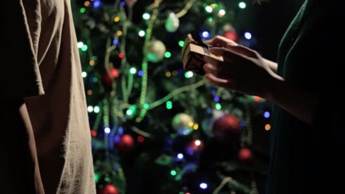 手中的圣诞礼物。穿着浅棕色t恤的男朋友给女孩穿绿色针织夹克，用老虎纸包裹，用黑色丝带绑在模糊新年树的