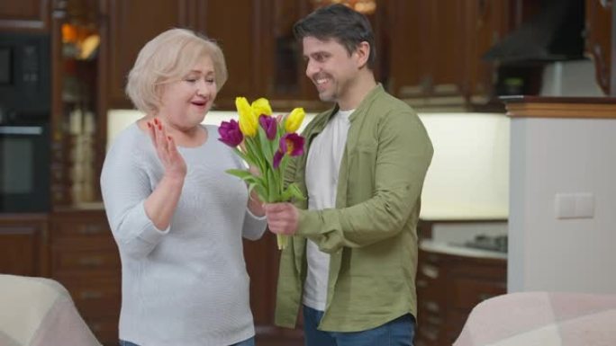 惊讶的高级美女拥抱成年男子，微笑着送一束鲜花。兴奋的高加索母亲退休人员的肖像感谢在室内拥抱儿子的关注