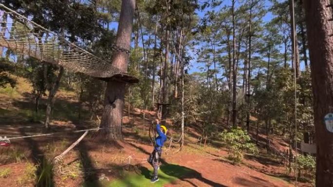 一个戴着黄色头盔的男孩的慢动作拍摄，该男孩在爬绳冒险公园里玩得很开心。在电话上拍摄