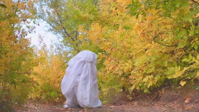 秋天森林里一个穿着幽灵服装的男人在下午庆祝万圣节。鬼魂鬼混，拍拍手，穿着白纸玩得开心