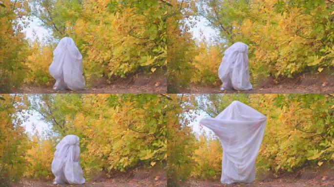 秋天森林里一个穿着幽灵服装的男人在下午庆祝万圣节。鬼魂鬼混，拍拍手，穿着白纸玩得开心