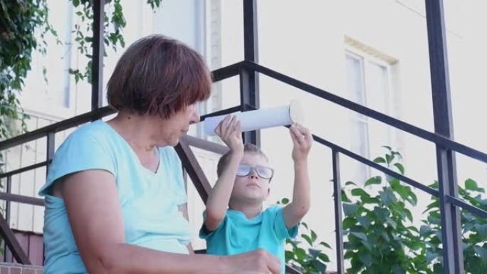 祖母和她的孙子男孩在房子后院的楼梯上欢乐地玩耍，在玩具纸管望远镜中探索星座。亲戚的交流和世代的交流