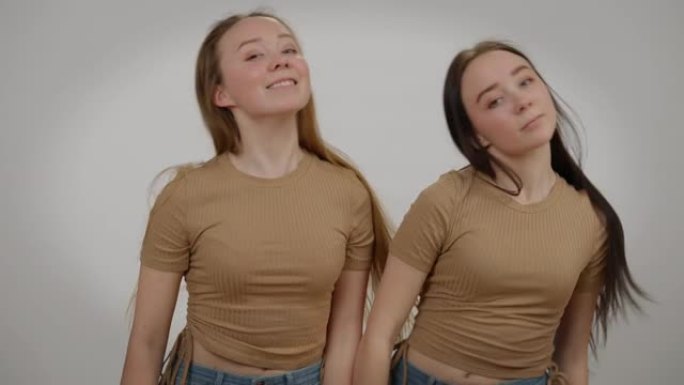 苗条美丽的同卵双胞胎姐妹同时在灰色背景下看着相机跳舞。前视图中拍摄的快乐迷人的高加索兄弟姐妹微笑的肖
