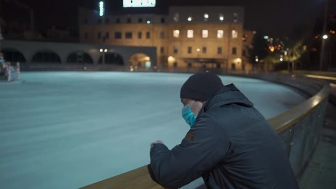 晚上，在冠状病毒大流行的禁闭期间，一名戴着面具的悲伤男子看着一个室内冰场，溜冰场中央有一棵圣诞树。空