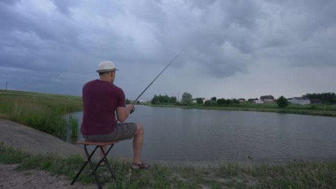 男子钓鱼坐在池塘岸上的椅子上，从后面看
